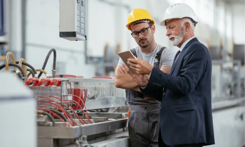 A senior project manager in a suit and hard hat reviewing digital plans on a tablet with a technician on a factory floor.