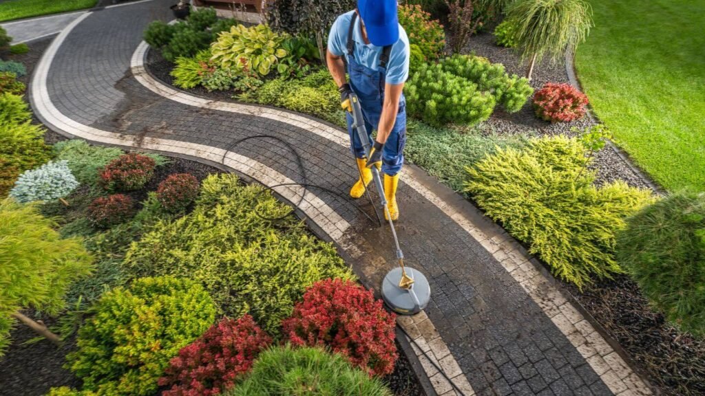 A professional technician using a surface cleaner to pressure wash a curved stone paver walkway surrounded by manicured landscaping.